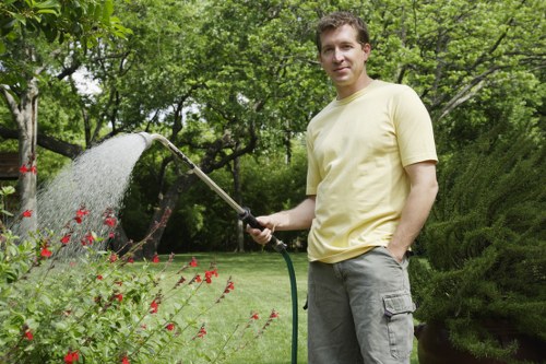 A man in a yellow t-shirt and grey trousers is standing in a well-maintained garden in Kings Cross, watering a flower bed with a garden hose. The flower bed contains red flowers, bordered by green foliage, and is situated adjacent to a lush, green lawn with dense grass that appears recently mown. Behind him, the garden features a background of tall trees and bushes with vibrant green leaves, creating a natural, shaded environment. The scene is outdoors on a partly cloudy day, with soft, natural lighting illuminating the garden space. The overall setting reflects a tidy, landscaped outdoor area typical of residential gardens in London postcodes, and is indicative of professional gardening and lawn care services provided by companies such as Garden Maintenance Kings Cross to maintain such a lush, inviting outdoor environment in the area.